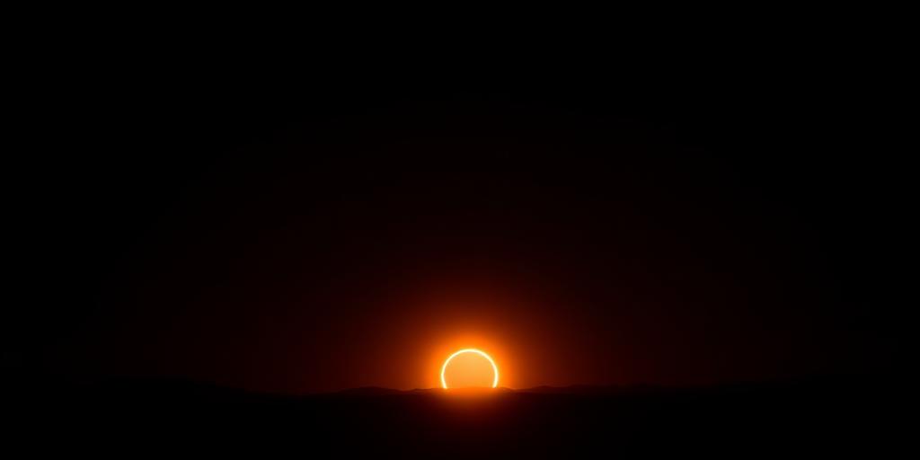 Eclipsed sun low over a glowing orange horizon with dark hills